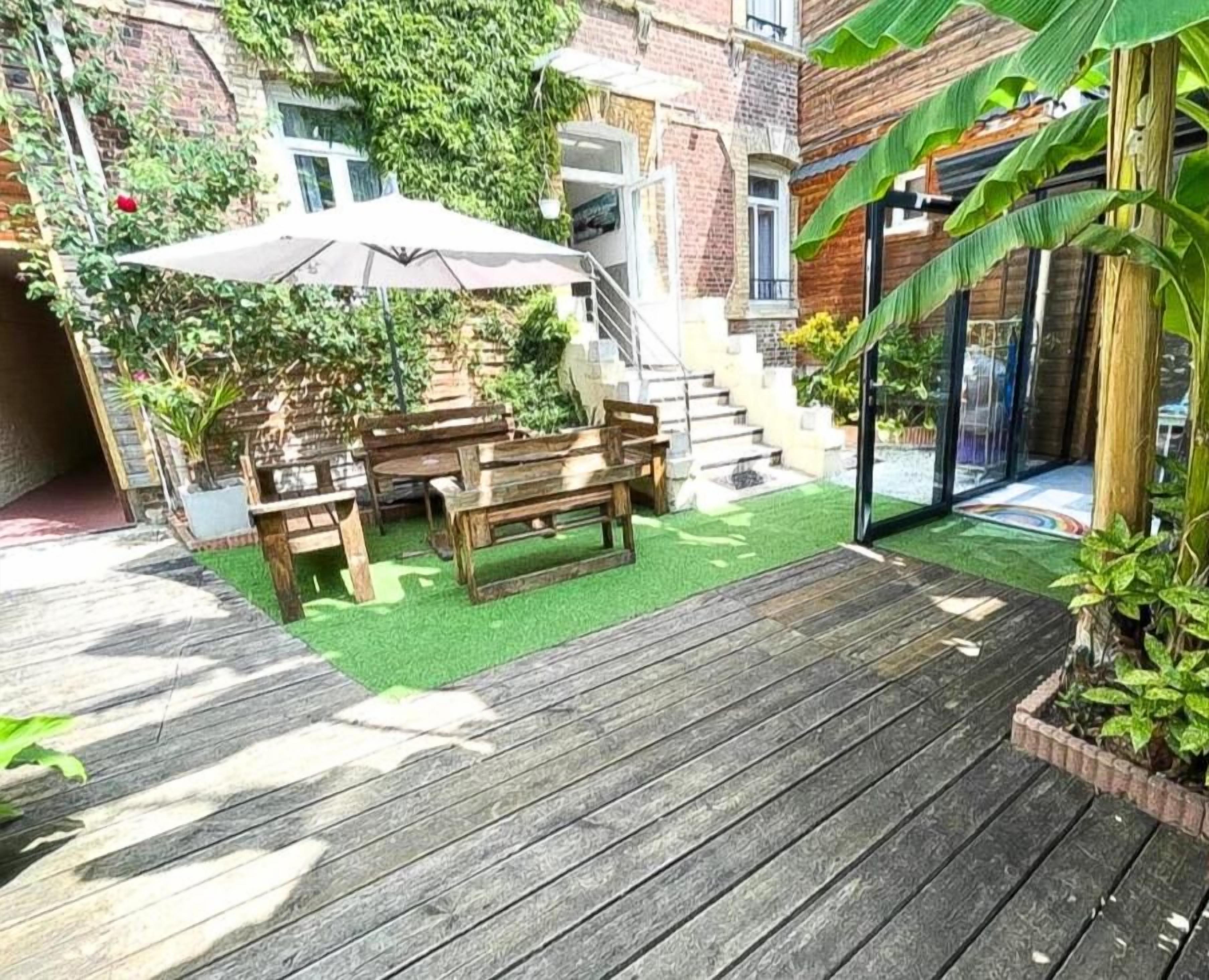 Een houten terras met een tafel en stoelen, omringd door groen en een bakstenen gebouw.