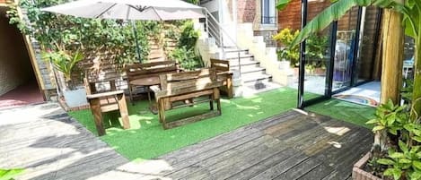 Um deck de madeira com mesa e cadeiras, rodeado por vegetação e um edifício de tijolos.
