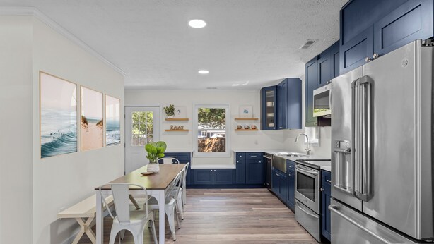 A modern kitchen with a wooden dining table, white chairs, and stainless steel appliances. - 4 min to beach, fully fenced yard + games, pet and kid friendly! (Oak Island)