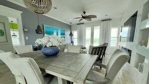 Un coin repas avec une table en bois, des chaises blanches, un vase bleu contenant des fleurs blanches, un lustre et un ventilateur de plafond.