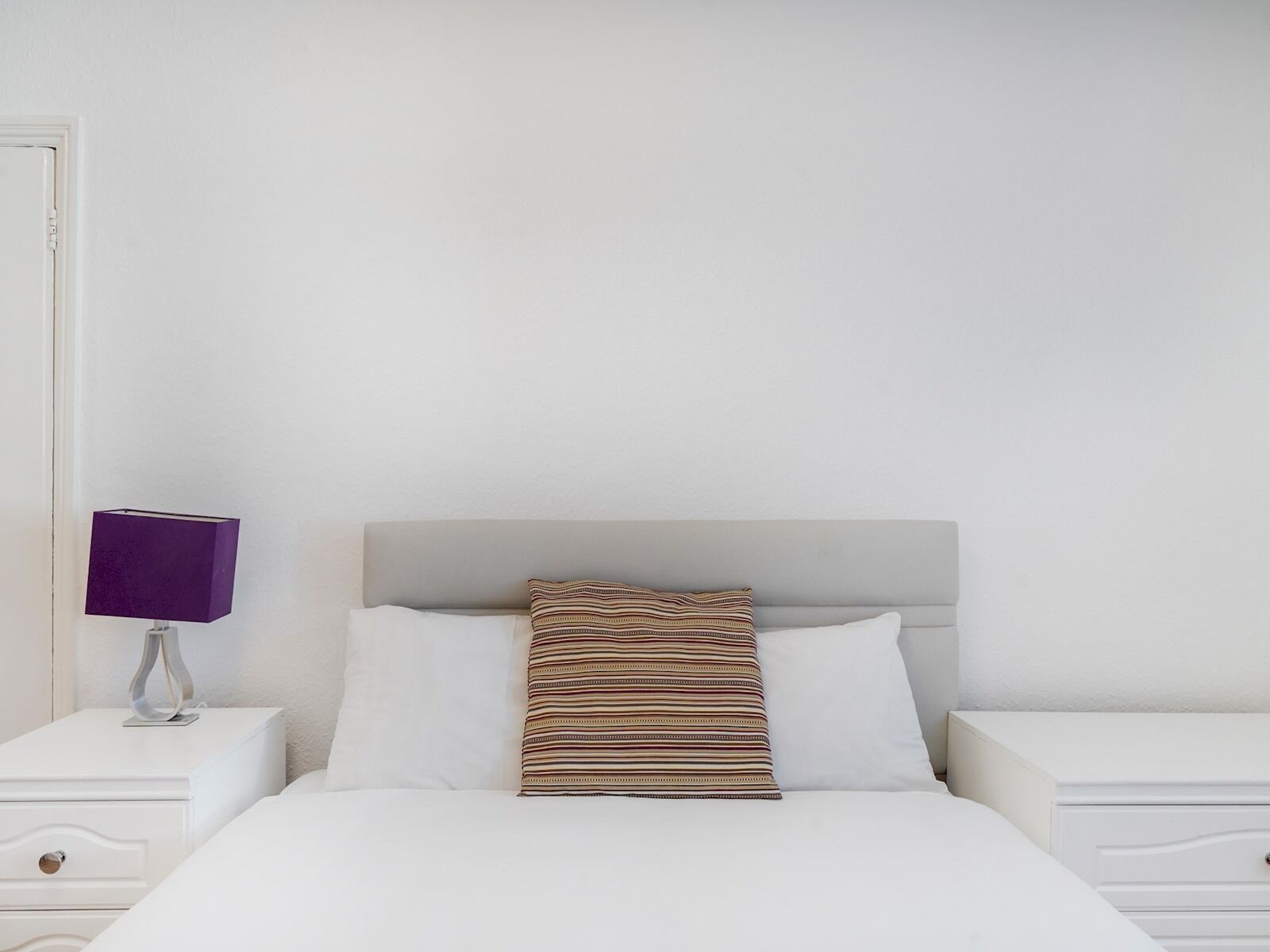 Een net opgemaakt bed met witte beddengoed en een gestreept kussen, een witte nachtkastje met een paarse lamp, en een witte dressoir met een lade.