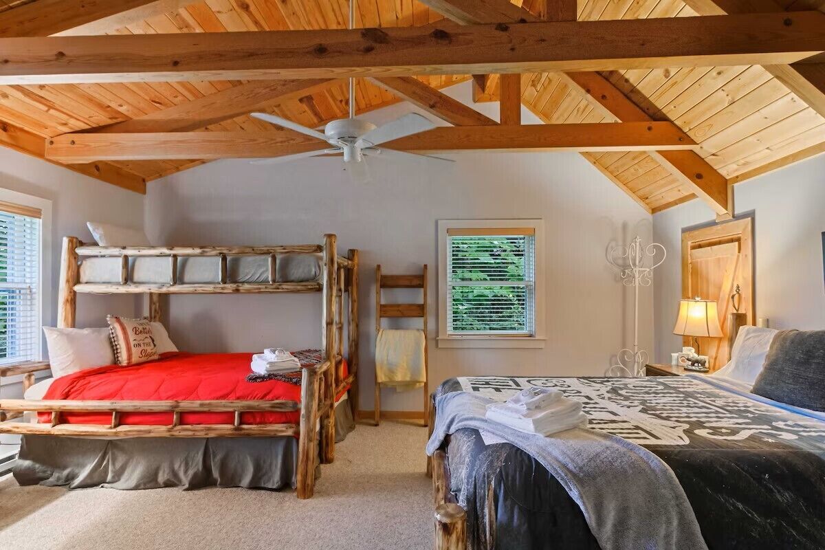 A room with a bunk bed, a single bed, a ceiling fan, a window with blinds, and a wall-mounted lamp.