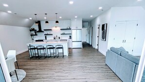 A spacious living area with a large wooden floor, a central island with bar stools, and a grey sofa.