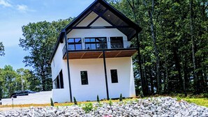 A modern two-story house with a balcony, surrounded by a rocky landscape and dense trees.