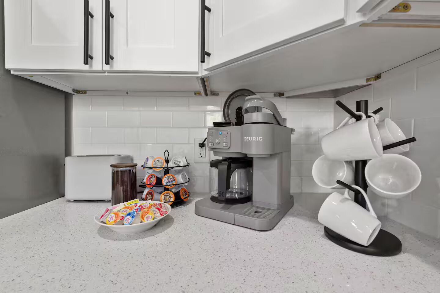 Une cuisine avec des meubles blancs, une machine à café Keurig, une autre machine à café et un plateau rempli de tasses colorées.