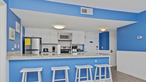 A modern kitchen with white cabinets, a granite countertop, and a blue accent wall. There are three white bar stools and a microwave above the counter. - Updated 2BR MBR Condo. Walk to Beach, Pools, Lazy River & Gated Resort (Myrtle Beach)