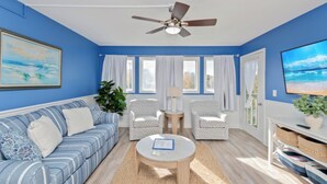 A living room with a blue wall, a ceiling fan, a sofa, two armchairs, a coffee table, and a TV. - Updated 2BR MBR Condo. Walk to Beach, Pools, Lazy River & Gated Resort (Myrtle Beach)