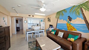 Ein Wohnzimmer mit braunem Ledersofa, einem Couchtisch, einem Esstisch mit Stühlen, einer Küchenzeile und einem Wandgemälde einer Frau am Strand.