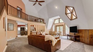 Een ruime woonkamer met een grote bruine bank, een houten bureau en een televisie. Er is een balkon met een hekwerk en een plafondventilator.