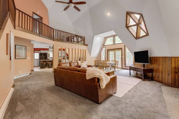 A spacious living area with a large brown sofa, a wooden desk, and a television. There is a balcony with a railing and a ceiling fan.