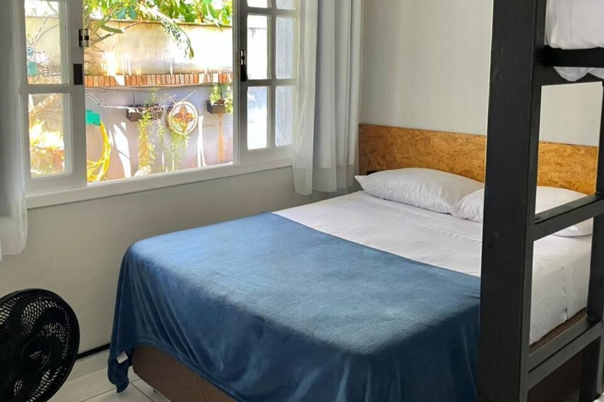 Een kamer met een bed, een raam met gordijnen, een ventilator en uitzicht op groen.