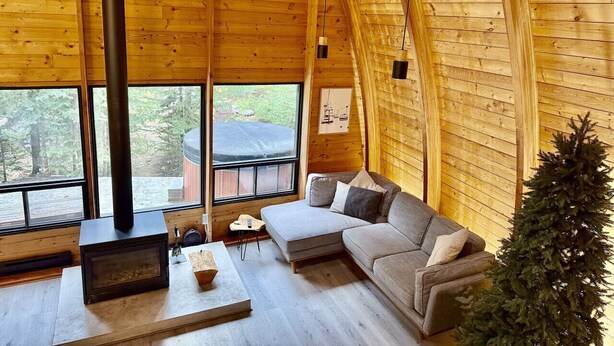 Interior acogedor de una cabaña con sofá, chimenea y ventanas amplias que ofrecen vistas al exterior.