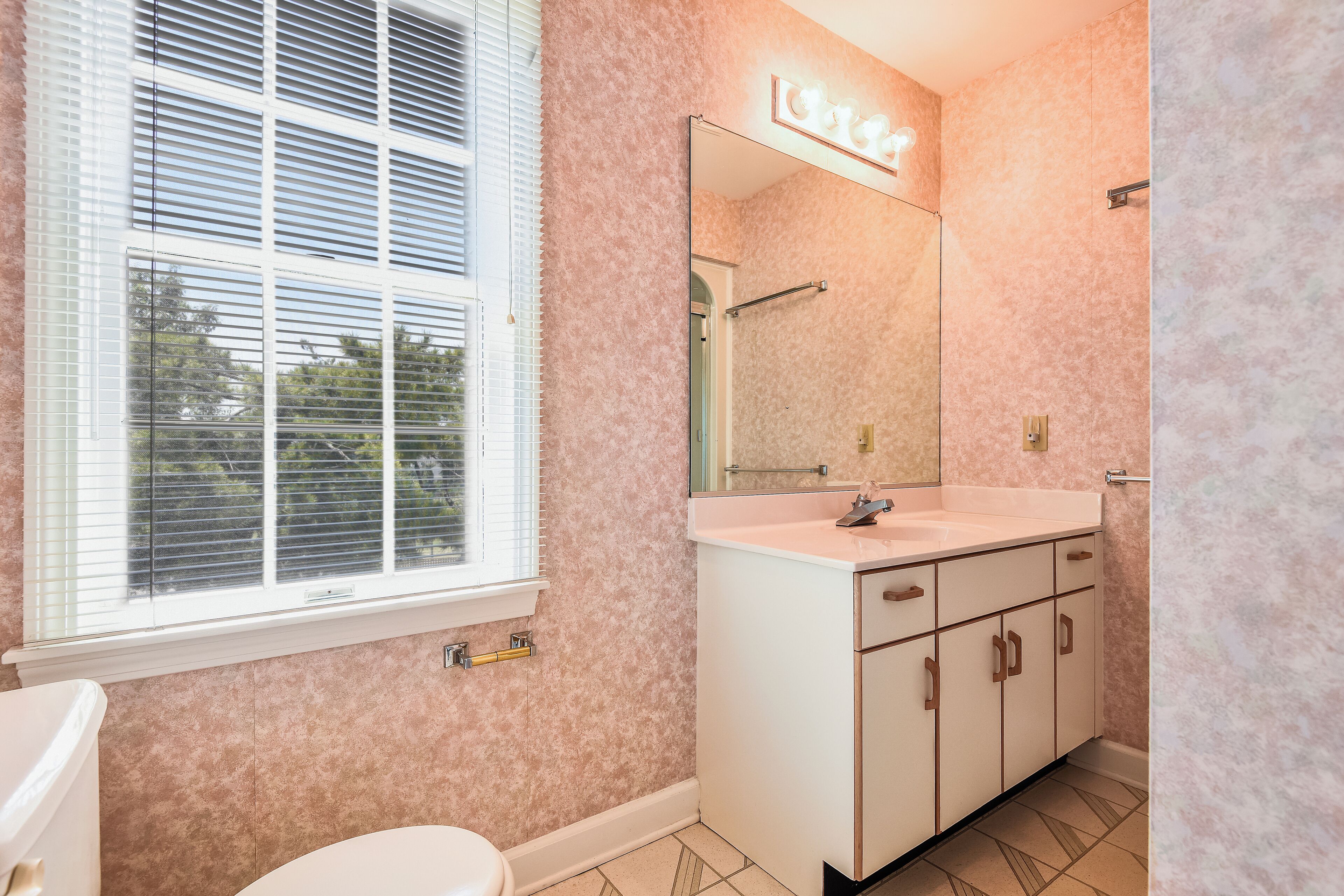 A bathroom with a white vanity, a mirror with lights, a window with blinds, and a toilet.