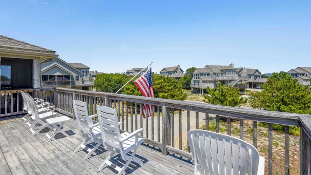 Une terrasse en bois avec des chaises blanches, un drapeau américain et, au loin, des maisons situées en bord de mer.