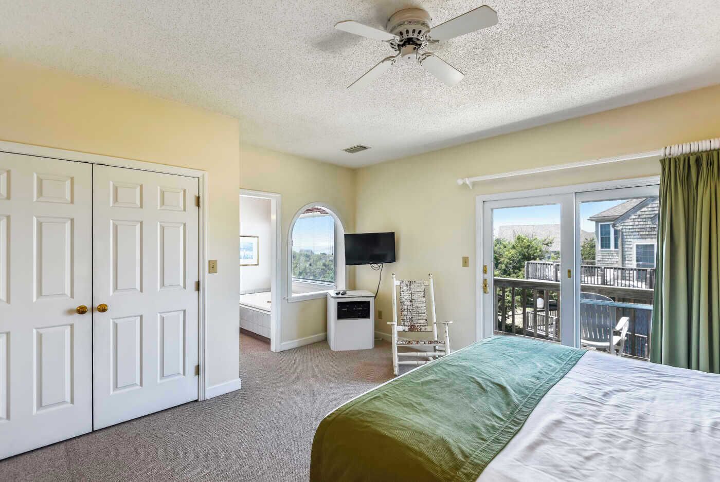 A bedroom with a bed, a TV, a desk, and a chair. There is a sliding glass door leading to a balcony.