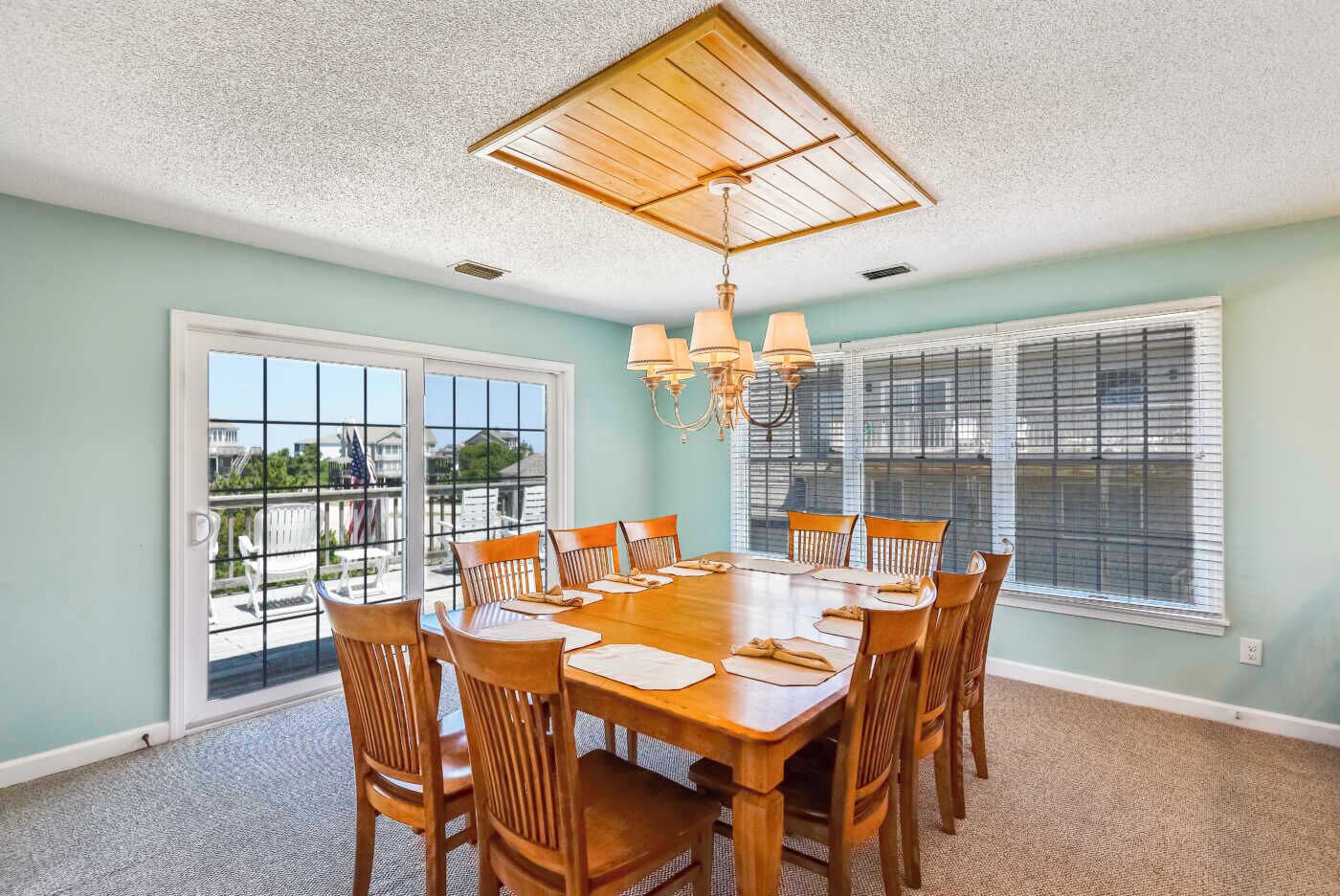 A dining room with a large wooden table and chairs, a chandelier, and large windows with blinds.