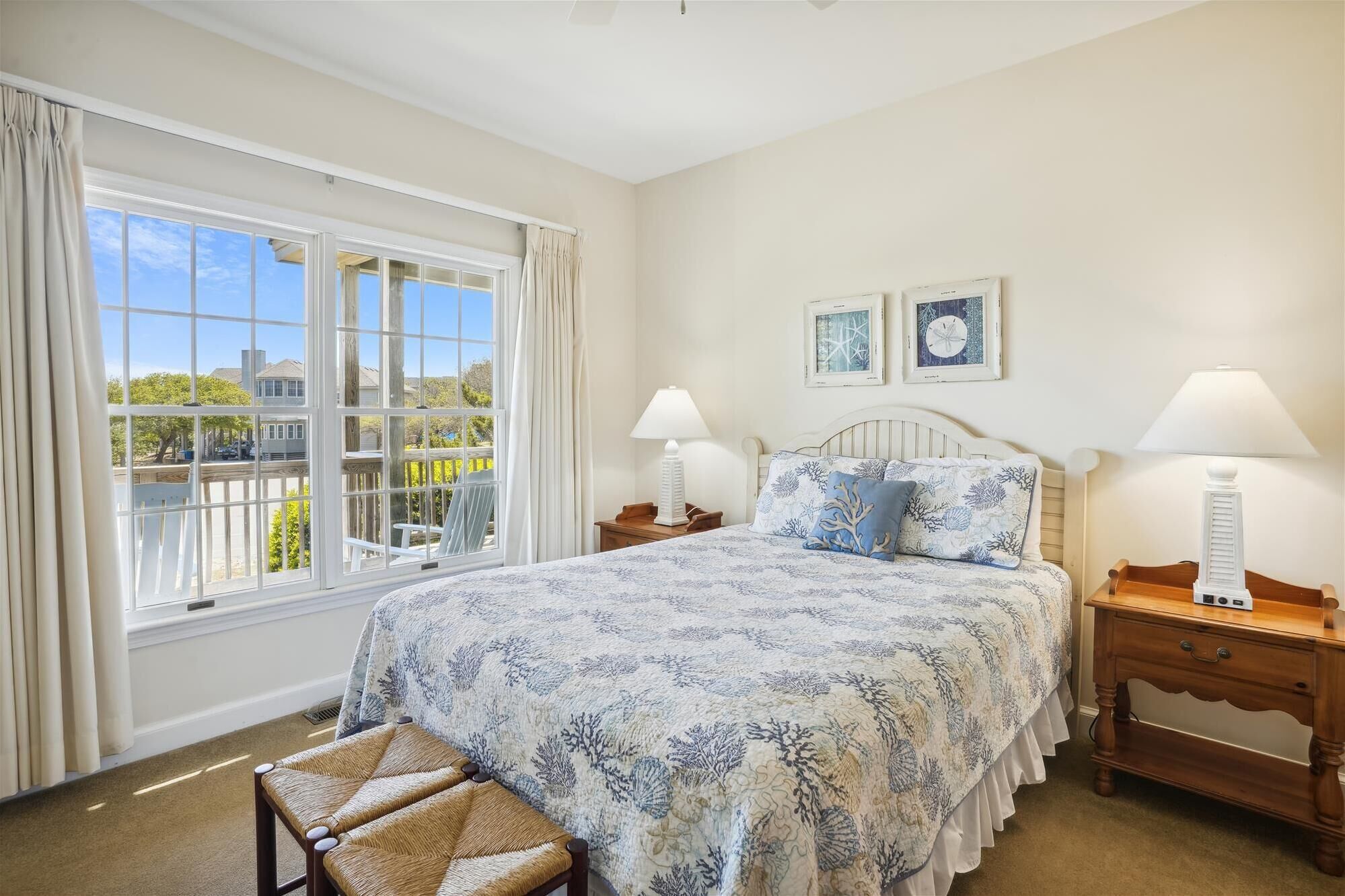 A bedroom with a large window, a bed with a floral bedspread, two bedside lamps, and a wooden nightstand.