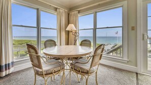 Una habitación con una mesa redonda y cuatro sillas, ventanas amplias con vista al océano y una lámpara sobre la mesa.