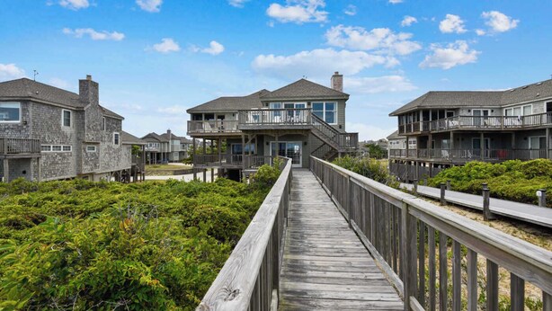 Un muelle de madera que conduce a una propiedad frente a la playa con casas de varios pisos, con balcones y ventanas grandes.