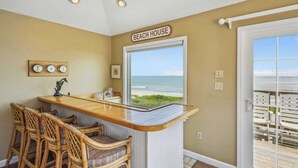 Interior de uma casa de praia com área de bar, cadeiras de vime, uma janela grande com vista para o oceano e uma placa com a inscrição 'BEACH HOUSE'.