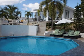 Un espace piscine moderne doté d’une piscine rectangulaire, de chaises longues et d’un escalier menant à une terrasse couverte.