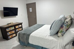 Une chambre à coucher avec un lit, un téléviseur fixé au mur, une porte et une commode en bois avec des tiroirs.