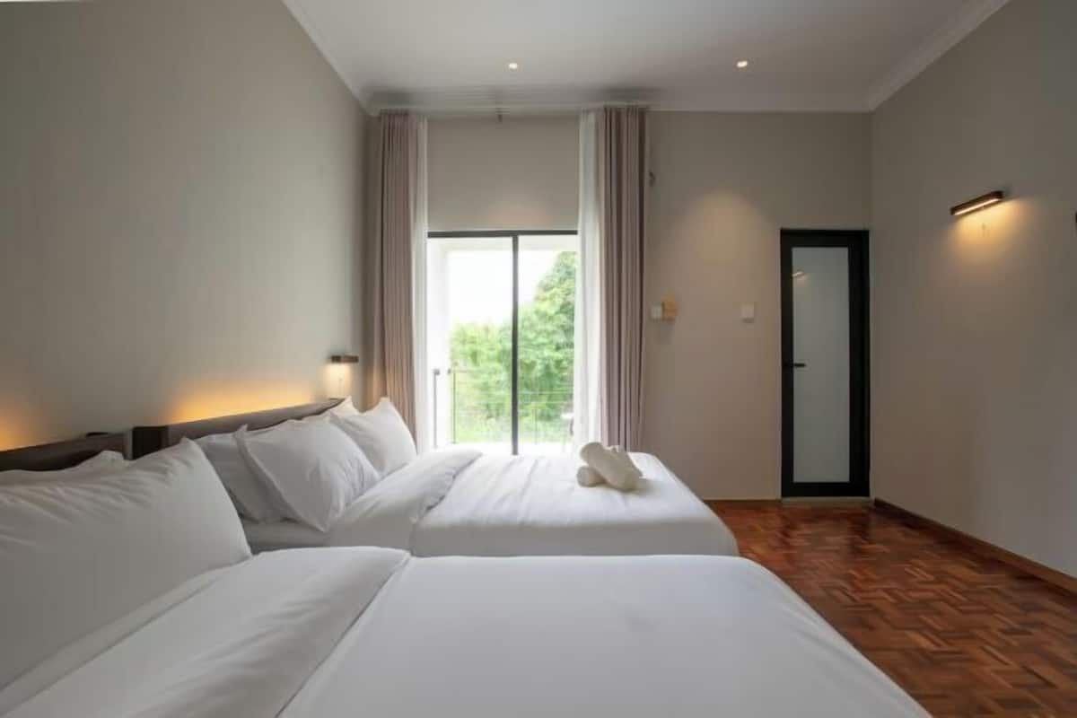 Une chambre d’hôtel avec deux lits, une grande fenêtre avec des rideaux, un plancher en bois et une porte à cadre noir.