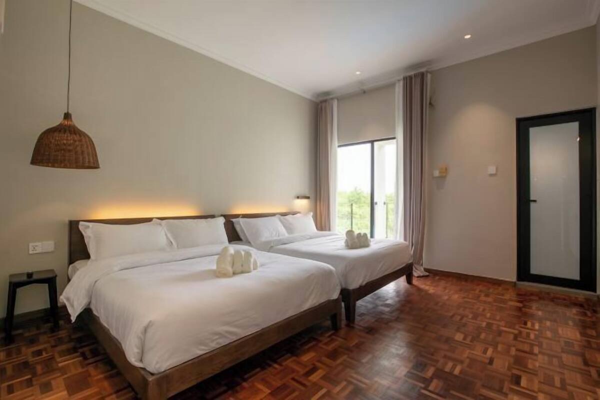 Une chambre d’hôtel avec deux lits, un plancher en bois, une grande fenêtre avec des rideaux et une lampe suspendue.