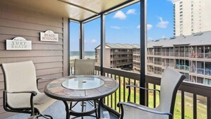 Een balkon met een glazen tafel en stoelen, borden die een strandbungalow en een surfhut aangeven, en een uitzicht op een gebouw met balkons.