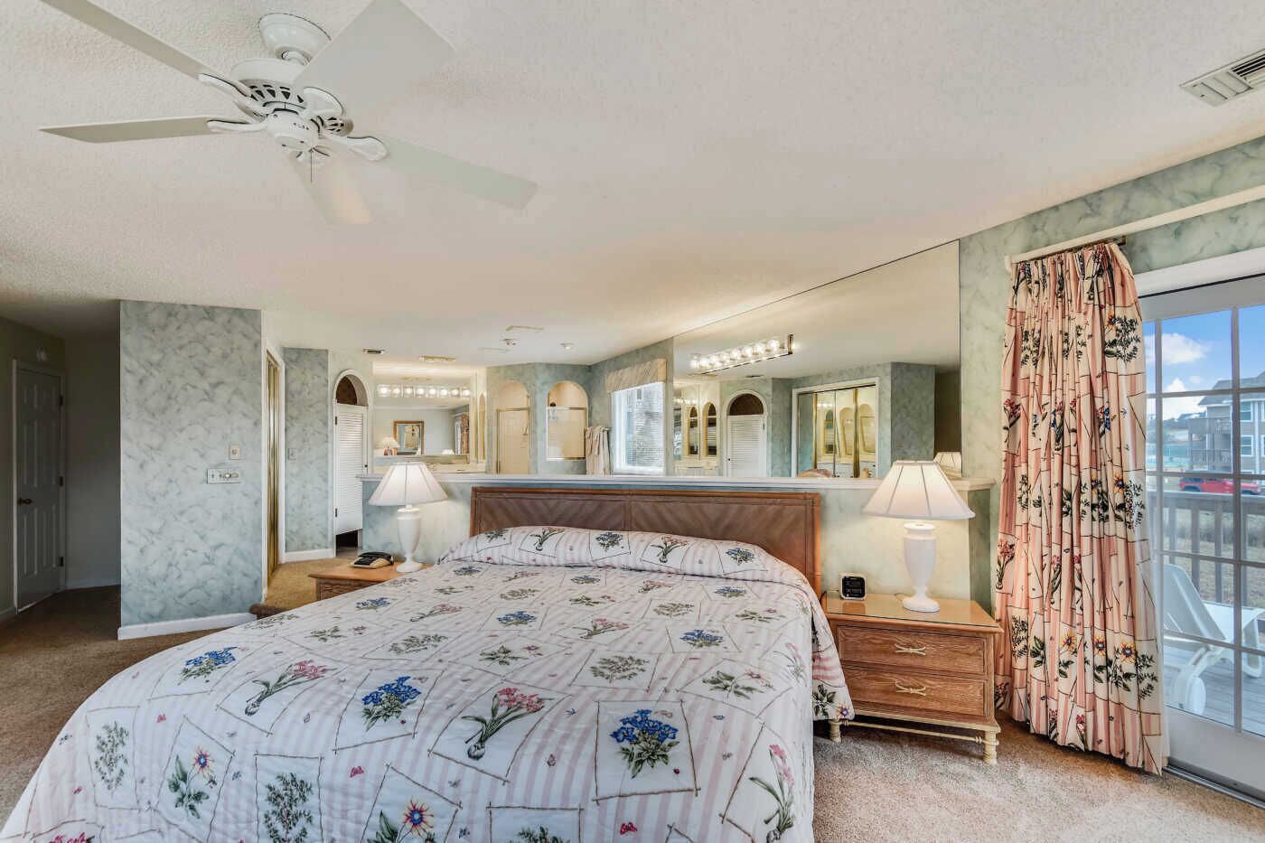 A bedroom with a large bed, a ceiling fan, a vanity with mirrors, and a view of the outside through a window.