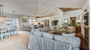 A spacious living area with a large grey sectional sofa, a dining table with chairs, a kitchen area with white cabinets, and a fireplace with a mirror above it. - Tappan, Ships Watch Community in Duck, NC (Duck)
