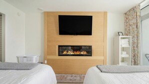 Une chambre d’hôtel avec deux lits, un téléviseur à écran plat fixé au-dessus d’une cheminée, et une fenêtre agrémentée de rideaux à motifs floraux.