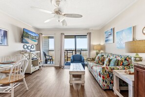 A room with a ceiling fan, a sofa with a tropical print, a dining table with chairs, and a view through a glass door. - Sea Cabin 120- Oceanfront 1BR/1BA- Pool (North Myrtle Beach)