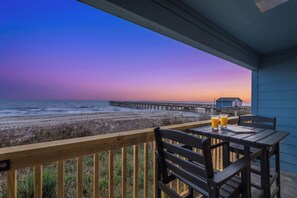 Une terrasse en bois avec une table et des chaises, donnant sur une plage et une jetée, au coucher du soleil.