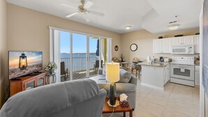 Uma sala de estar com televisão, sofá e uma mesa com luminária. Há uma varanda com vista para o mar.