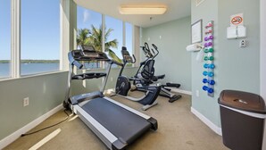 Uma sala de ginástica com esteira, elíptica e bicicleta ergométrica, suporte de halteres na parede e vista para a água.