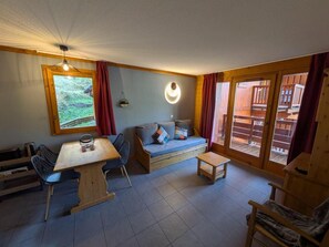 A cozy room with a wooden sofa, a dining table, and chairs. There is a window with curtains, a wall-mounted light fixture, and a view of an outdoor deck.