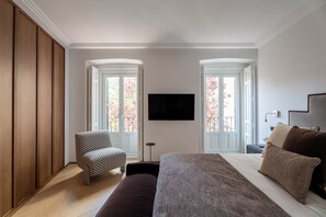 A bedroom with a bed, a chair, a TV, and a view of trees through the windows.