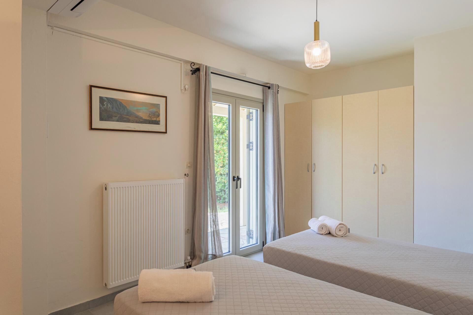 Une chambre avec deux lits, un radiateur, un tableau encadré, une armoire et une porte-fenêtre coulissante donnant sur l’extérieur.