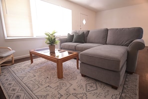 Un salon avec un canapé gris, une table basse en bois ornée d’une plante et des tapis à motifs.