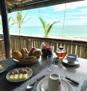 A table set with bread, pastries, a teapot, and a cup, overlooking a beach with palm trees. - Pousada San Antônio Praia  (Porto Seguro)