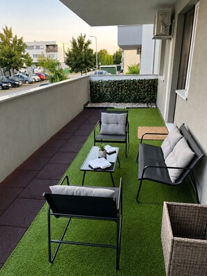A rooftop terrace with a green artificial turf, two black lounge chairs, a small table with a plant, and a wicker basket.
