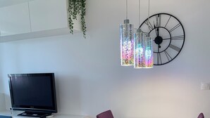 Un comedor moderno con una mesa blanca, sillas moradas y azules, un gran reloj de pared y un televisor sobre un mueble azul.