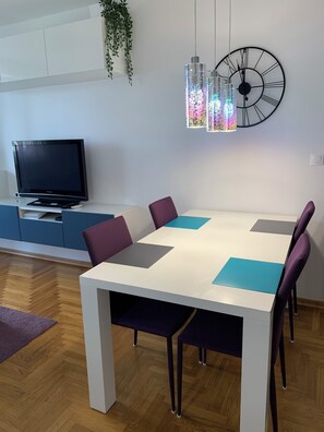 Un comedor moderno con una mesa blanca, sillas moradas y azules, un gran reloj de pared y un televisor sobre un mueble azul.