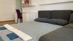Una cama bien hecha con un edredón estampado, una toalla blanca doblada y una toalla azul. Hay un sofá gris con almohadones, un escritorio blanco con una silla y un cuadro en la pared.