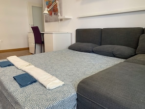 Una cama bien hecha con un edredón estampado, una toalla blanca doblada y una toalla azul. Hay un sofá gris con almohadones, un escritorio blanco con una silla y un cuadro en la pared.