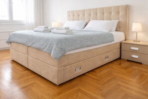 A neatly made bed with a tufted headboard, a bedside table with a lamp, and a wooden floor.
