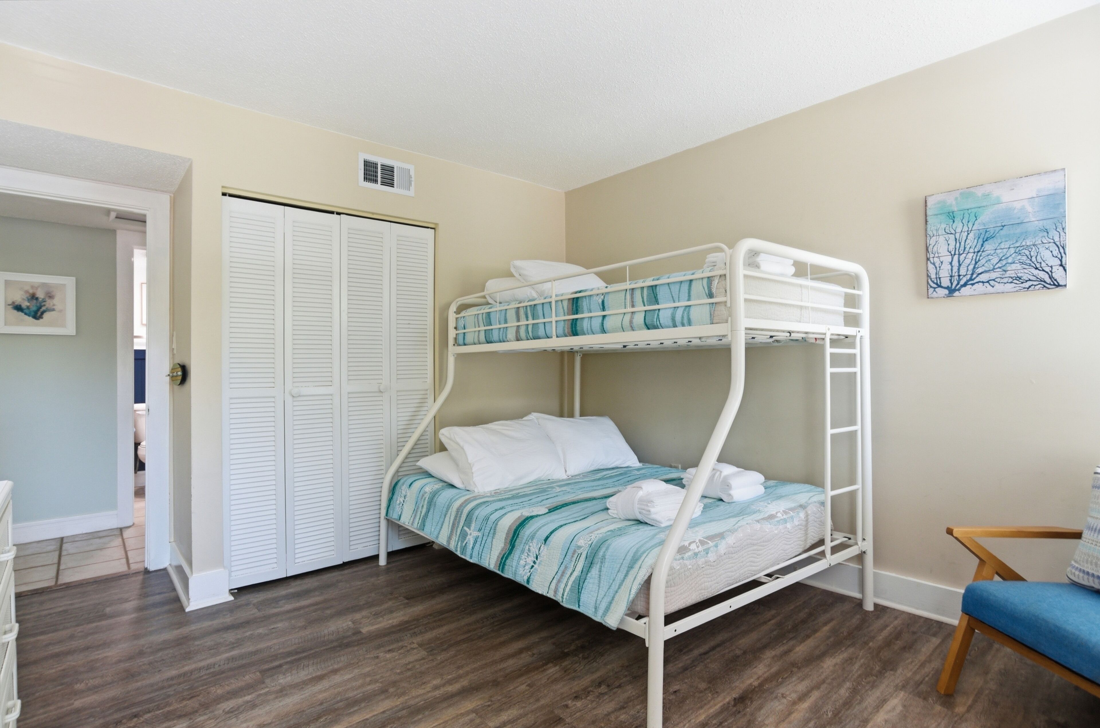 Ein Stockbett mit Streifenmuster, ein weißer Kleiderschrank mit Klappläden und ein blauer Stuhl.