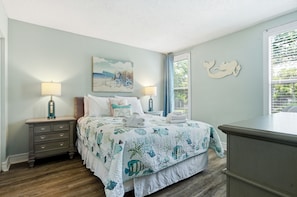 A bedroom with a bed, two bedside tables, a nightstand, a dresser, a window with blinds, and a wall with a painting and a decorative item.