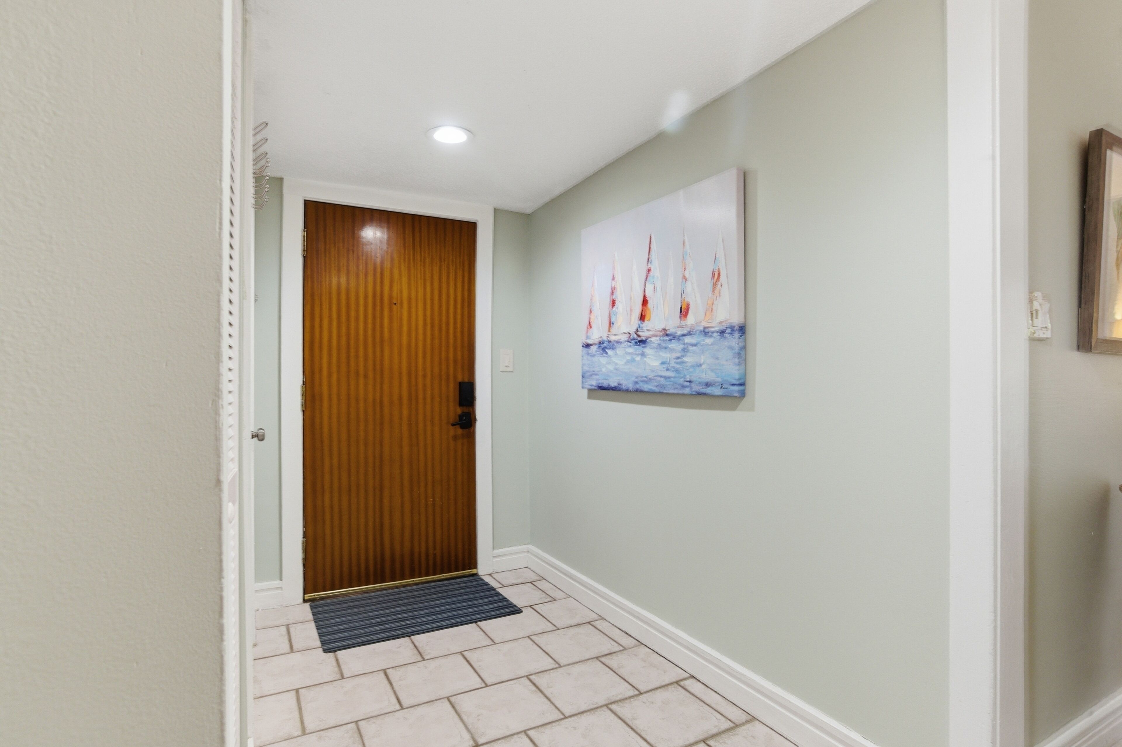 Un couloir avec une porte en bois, un tableau représentant des voiliers et un sol carrelé.